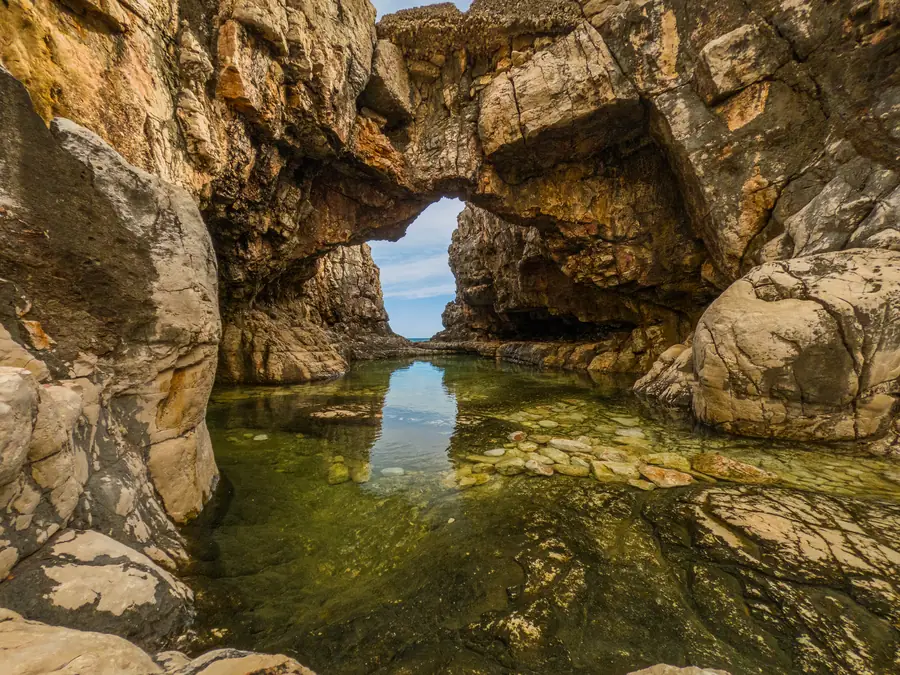 A sea arch on Lokrum island: eroded limestone framing a shallow tidal pool with pebbles visible through the water