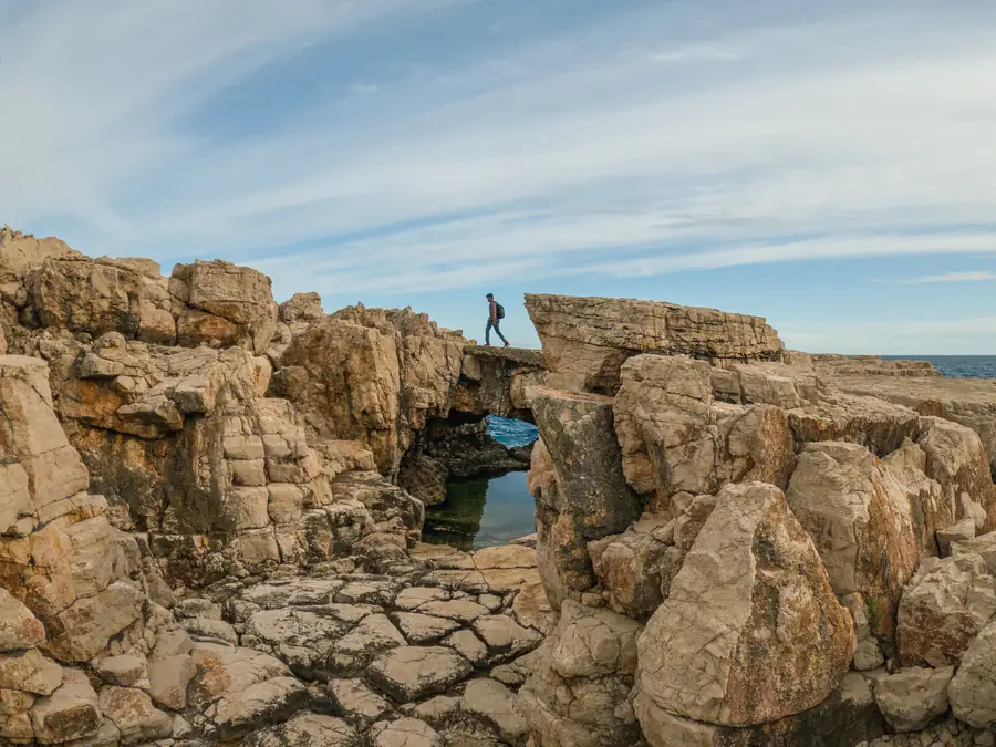A walker crossing a natural limestone bridge on Lokrum, with a tidal pool below and the open sea to the right