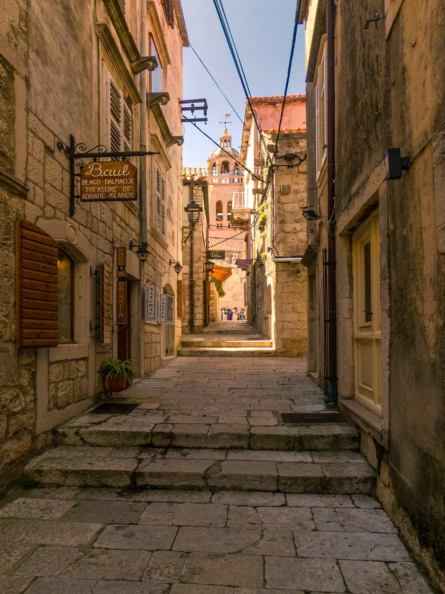 A stepped stone lane in Korcula old town leading to the cathedral bell tower, with shop signs and shuttered windows
