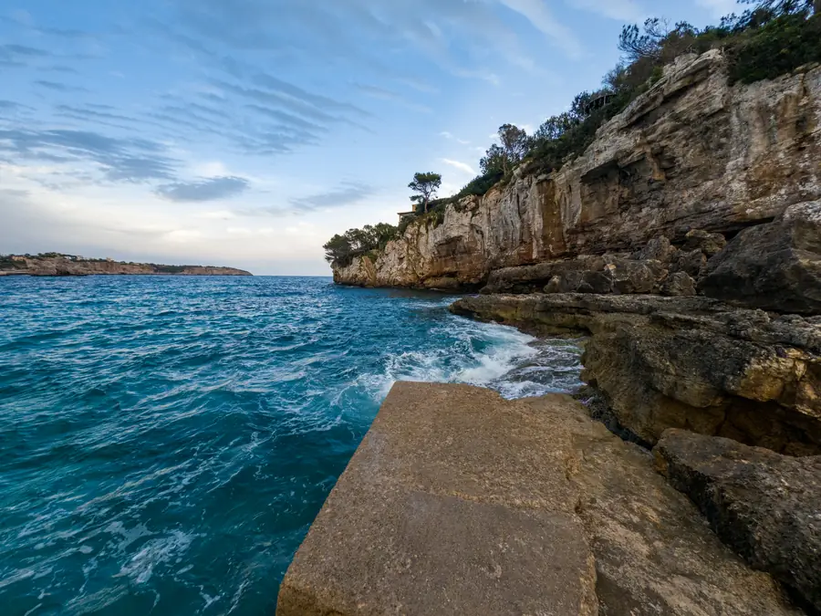 A limestone cove on the Mallorcan coast at dusk: turquoise water breaking against a rock shelf, with pines on the cliff above
