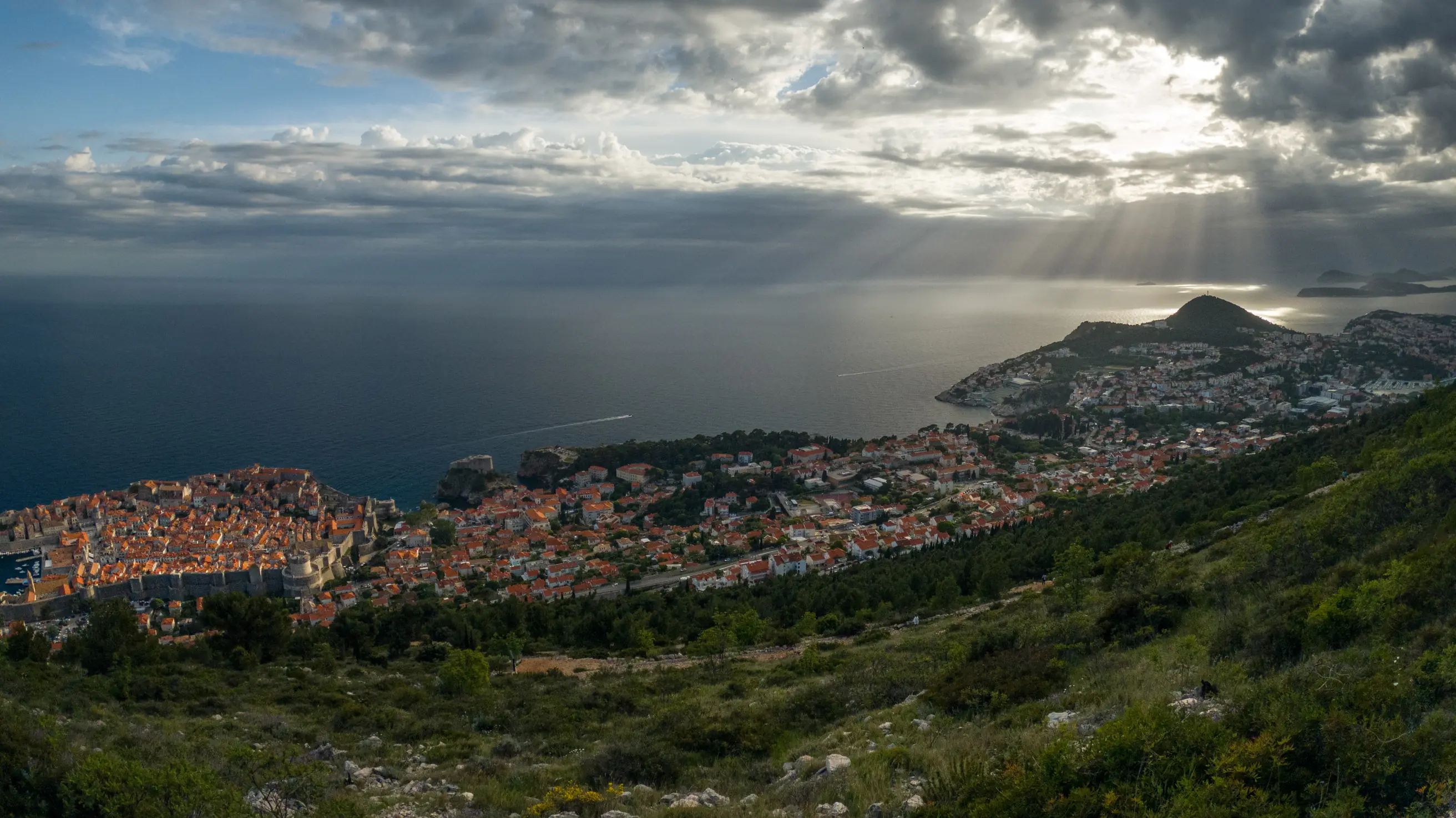 Dubrovnik from the hillside above: the walled old town on its promontory, sunbeams breaking through cloud over the Adriatic
