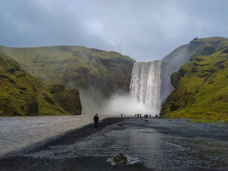 Skogafoss under a low grey sky: the falls dropping between mossy green slopes, with people small on the black gravel below