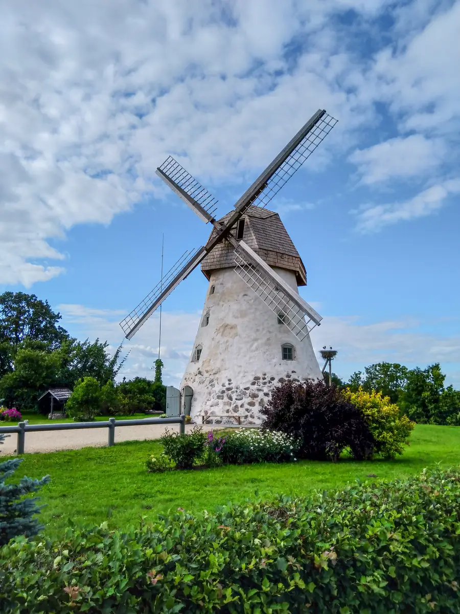 The windmill at Araisi: a whitewashed stone tower with a shingled cap and four lattice sails, above a clipped lawn and flowering shrubs