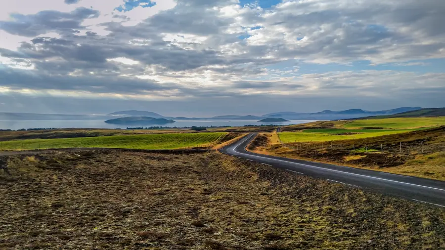 A two-lane road curving through Icelandic moorland toward a fjord, with low islands and hills under a broken sky