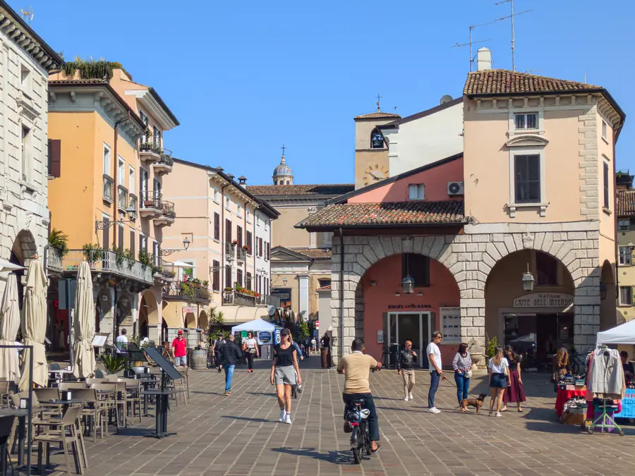 A sunlit square in Desenzano del Garda: arcaded stone buildings around a paved piazza, cafe tables in the shade and people crossing