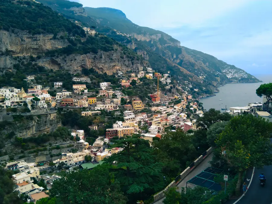 Positano from the road above: pastel houses stacked down a steep cliffside to the sea, with pines in the foreground