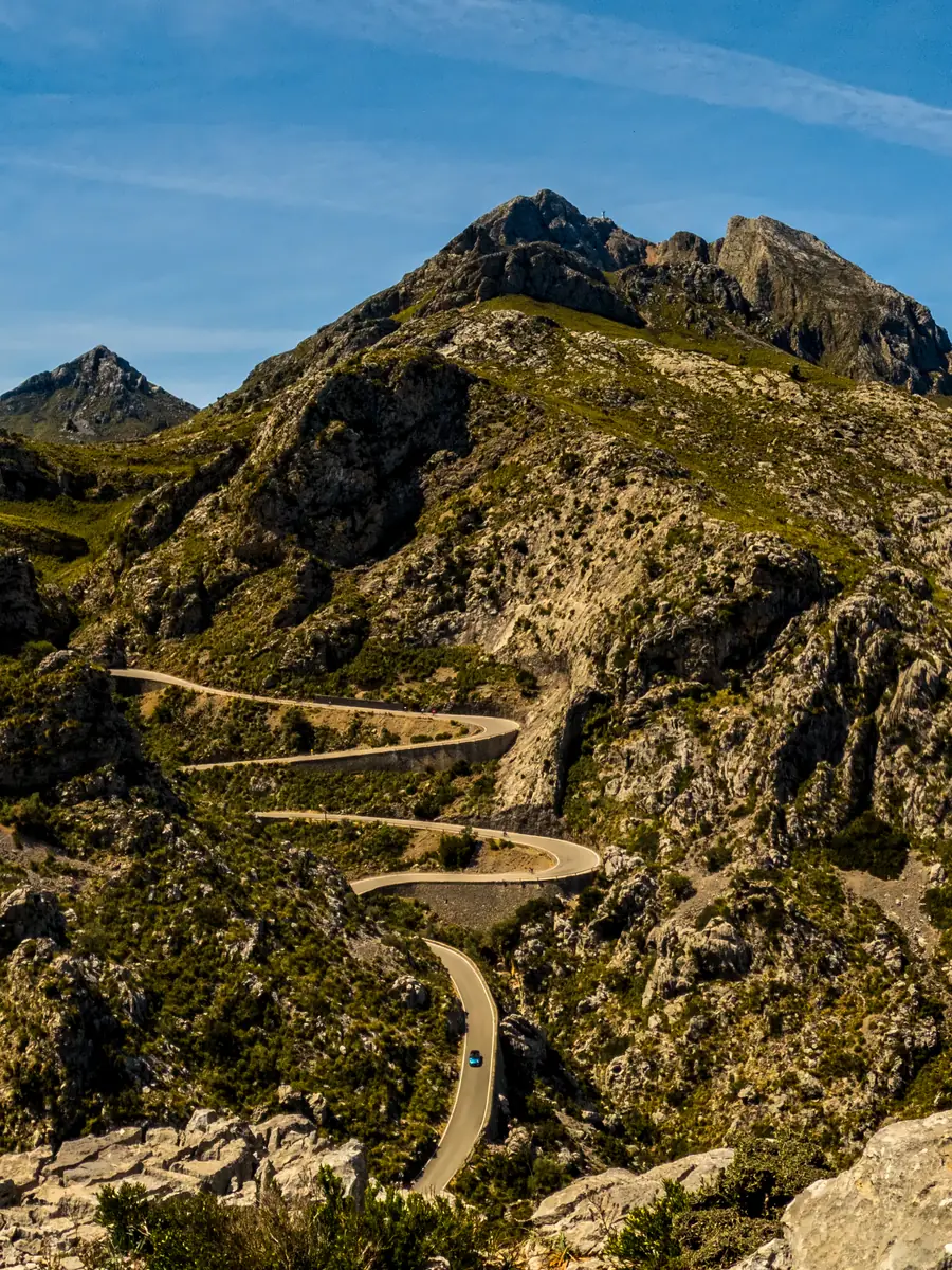 A hairpin road winding down through the Tramuntana mountains on Mallorca, a single car on the lower bend beneath a rocky summit