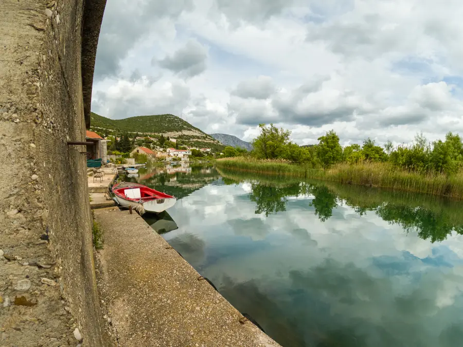 A red rowing boat moored along a concrete quay on the Neretva delta at Rogotin, clouds mirrored in the still channel