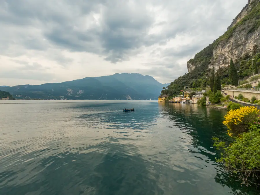 Lake Garda from the shore under heavy cloud: a small boat on flat water, cliffs and a lakeside hamlet on the right