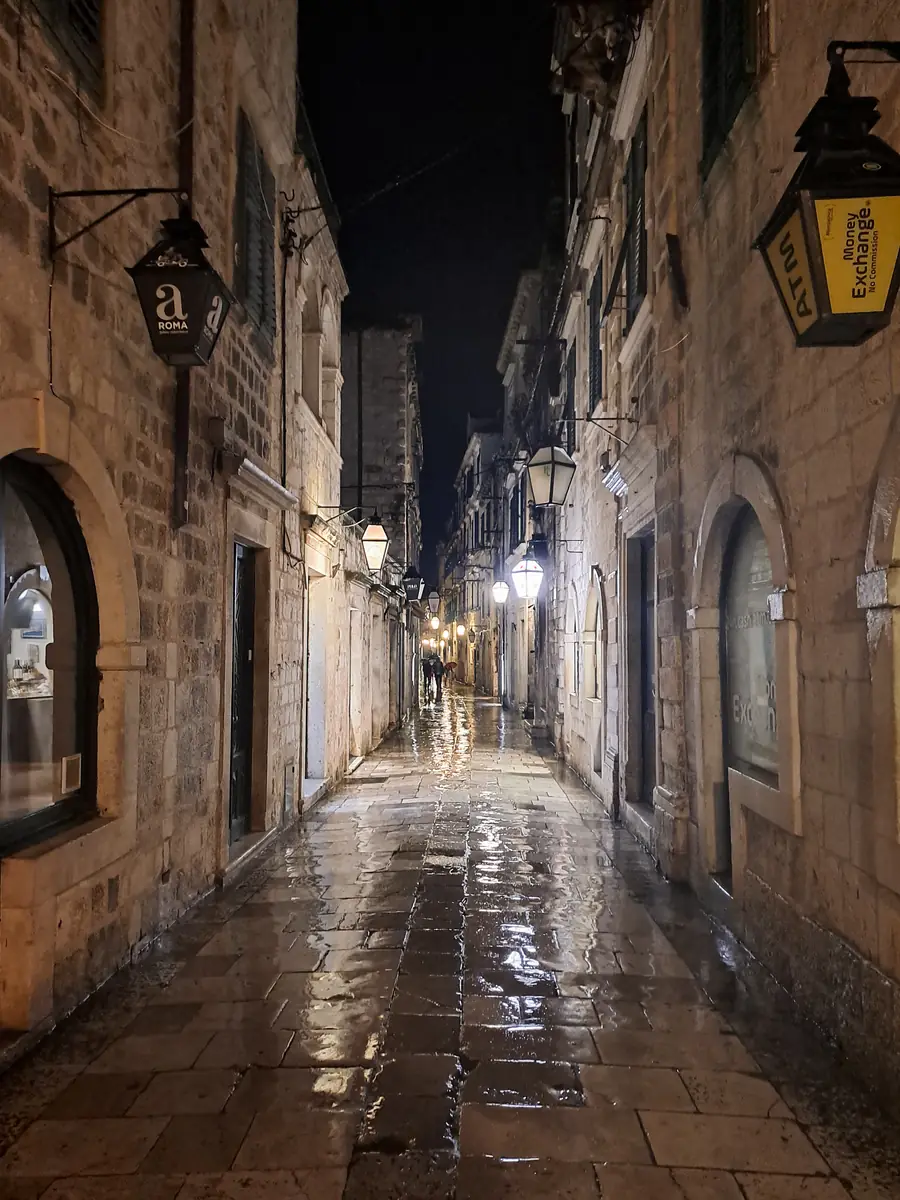 A narrow stone street in Dubrovnik's old town after rain at night, lanterns reflected in the wet paving and two figures far down the lane
