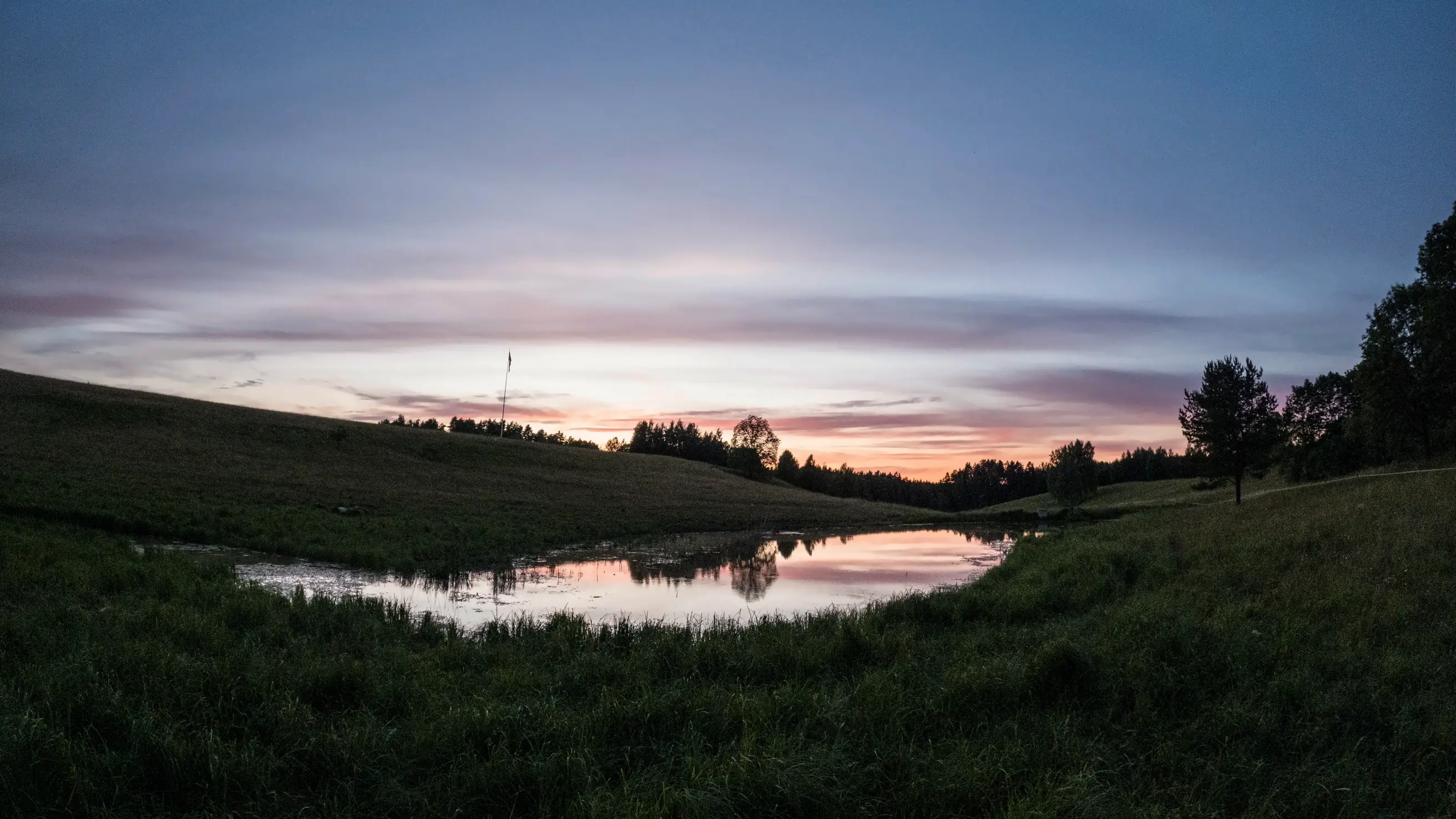 Dusk over a farm pond near Ligatne: the last of the sunset reflected in still water, grass slopes and a treeline in silhouette