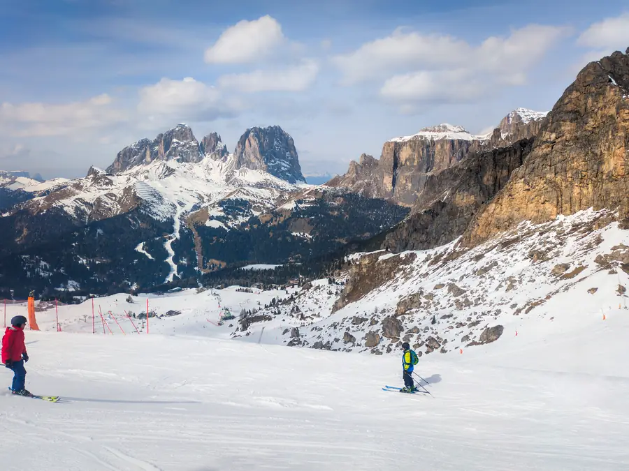 A groomed piste in the Dolomites: two skiers on the slope, with the Sassolungo massif and pale rock walls across the valley
