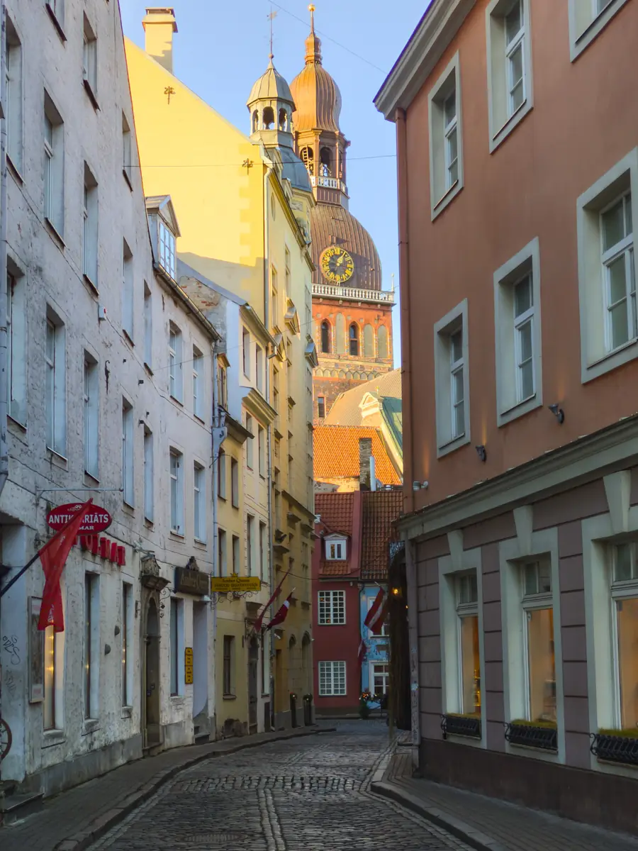 A cobbled street in Riga's old town at low sun, pastel facades on both sides and the cathedral tower rising at the end